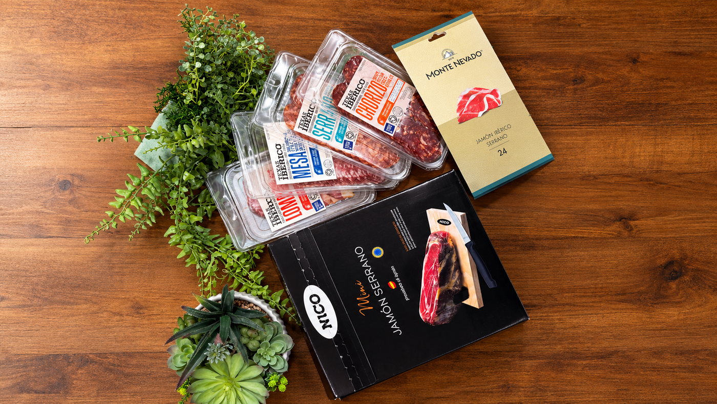 A curated food gift set arranged on a wooden tabletop, including several sliced cured meats, texas iberico lomo, texas iberico chorizo, texas iberico serrano salami slices, Mini serrano jamon, Monte Nevado iberico serrano jamon.