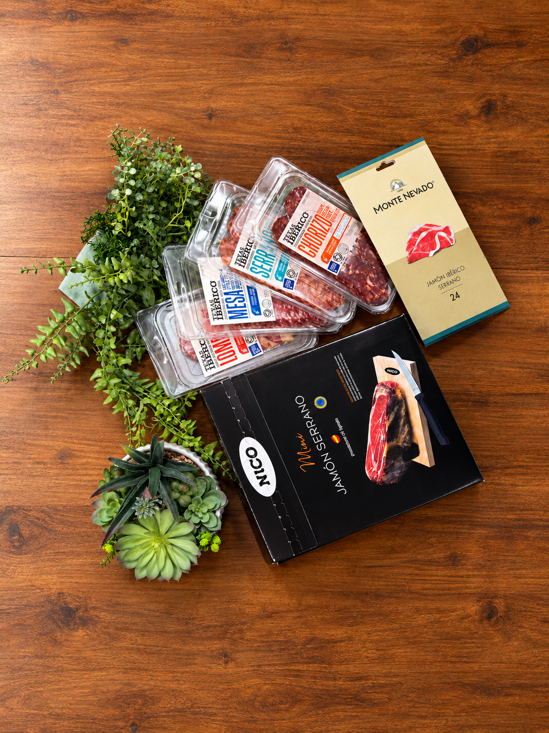 A curated food gift set arranged on a wooden tabletop, including several sliced cured meats, texas iberico lomo, texas iberico chorizo, texas iberico serrano salami slices, Mini serrano jamon, Monte Nevado iberico serrano jamon.